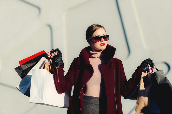 A woman outdoors, wearing an expensive dark red coat and sunglasses, carrying several shopping bags in both hands